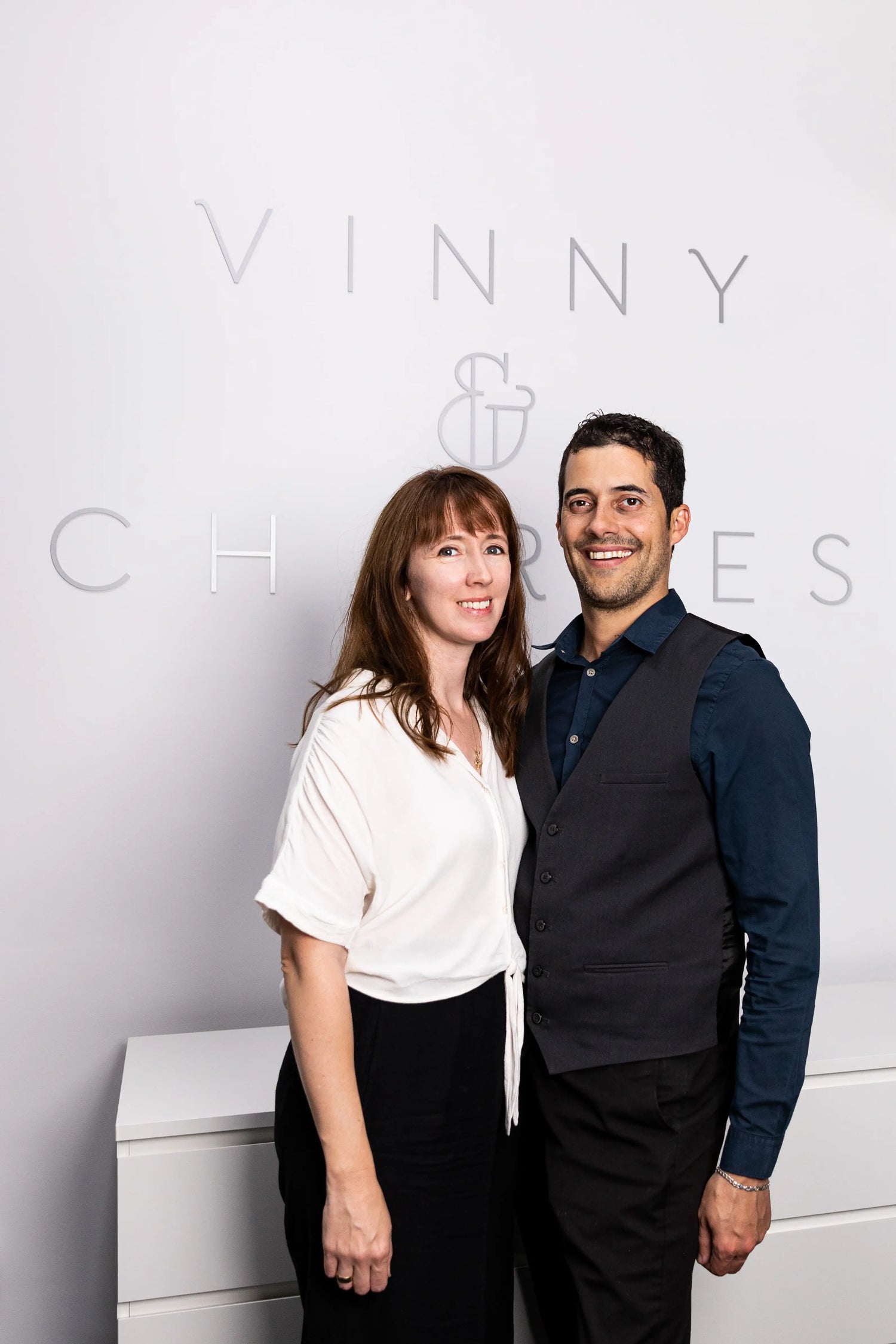 Photograph of Melody and Chris standing together, with the Vinny & Charles logo featured on the wall behind them. 